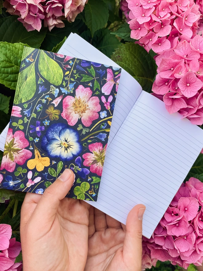 Hands Holding Two Small Botanical Print Notebooks with Pretty Pressed Flower Design on Pink Hydrangea Background