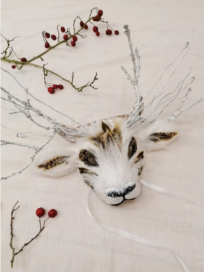 A white luxury deer part mask laying flat on a white cloth next to dried twigs