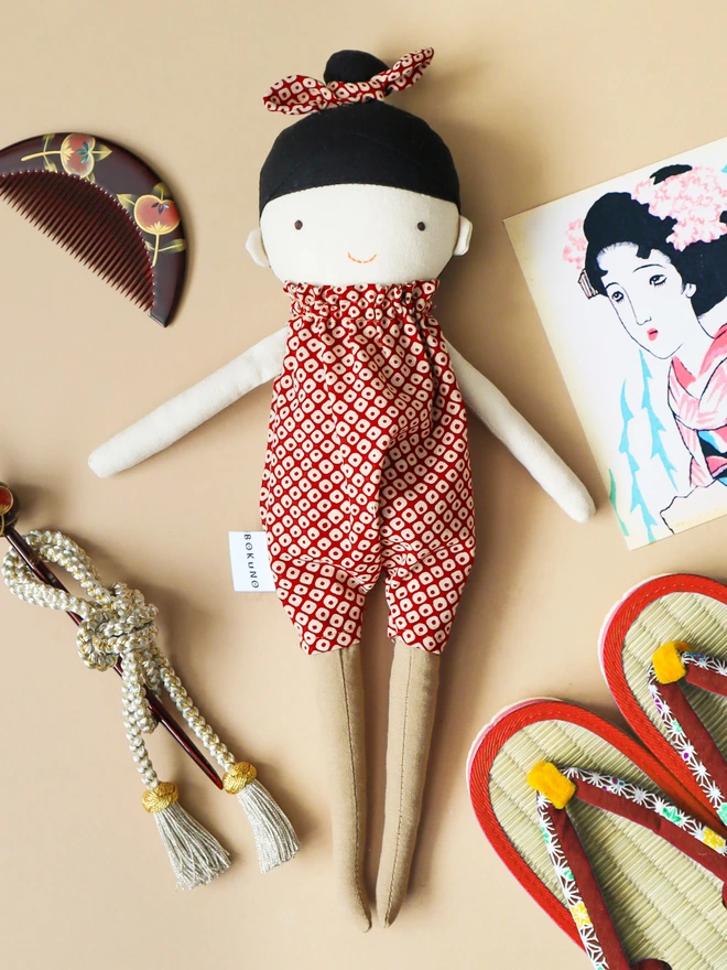 A handmade doll wearing a red and white Japanese shibori patterned jumpsuit with a matching bow in her black hair. The doll is surrounded by Japanese-inspired accessories, including a comb, hair stick, and sandals.