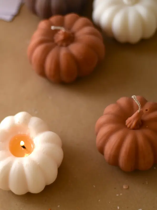 Medium Rust Wax Pumpkin Candle. A lit cream pumpkin candle glowing beside unlit terracotta pumpkin candles on kraft paper. The sculpted wax pumpkins bring warm, flickering light to autumn evenings.
