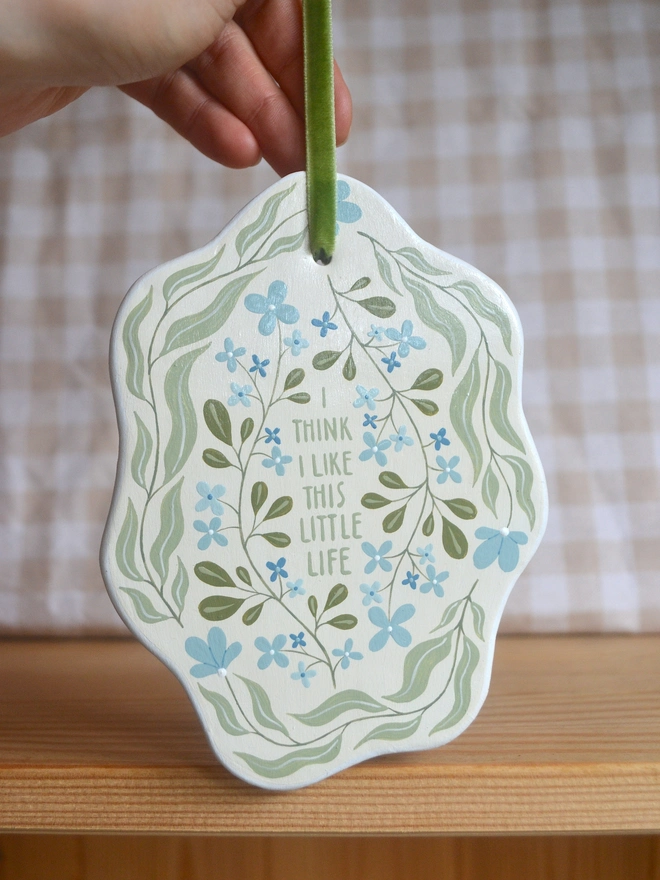 hanging clay sign with blue and green floral design reading 'i think i like this little life' in front of gingham background
