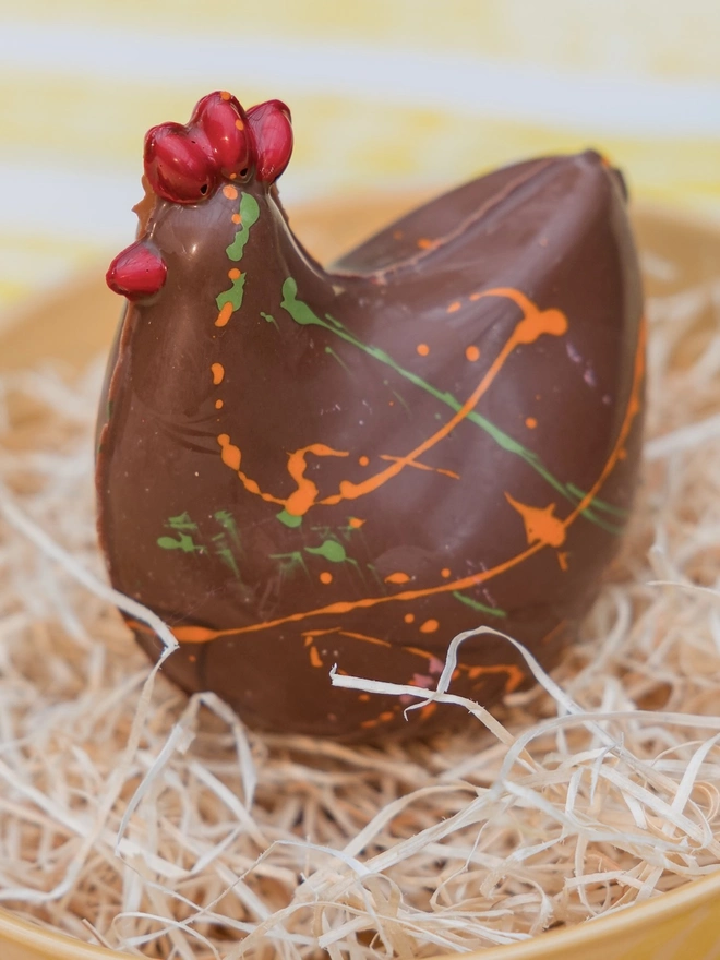 A milk chocolate Easter chicken from Nenette Chocolates sitting in  straw bedding in a yellow bowl, on a striped yellow and white tablecloth