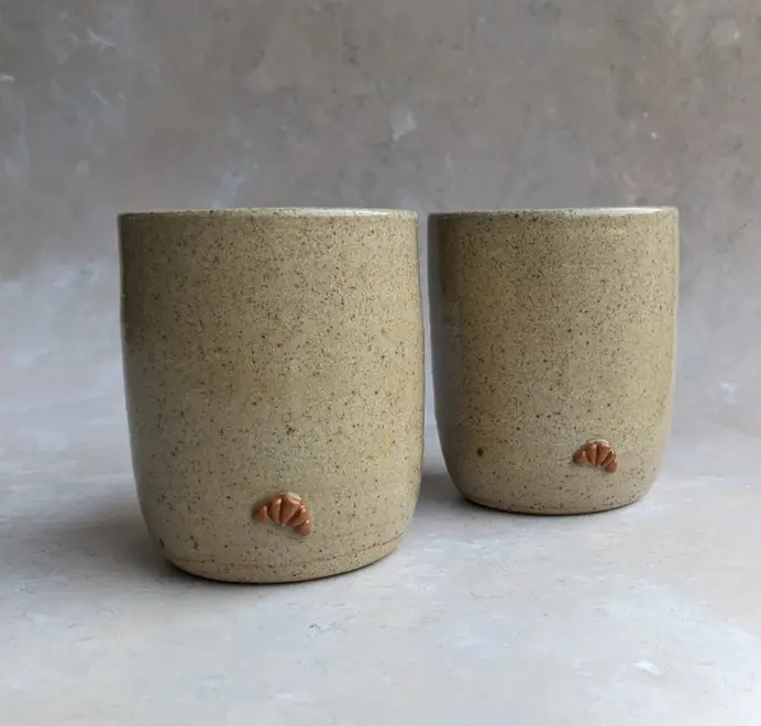 Two biscuit coloured croissant coffee cups with a subtle croissant design, placed against a grey marble background. 