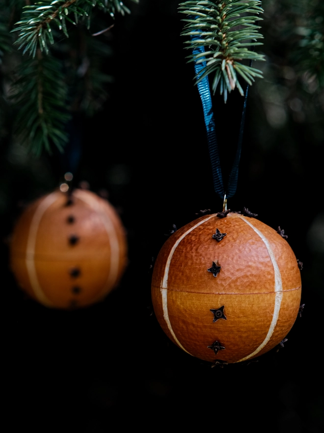 Handcrafted orange and clove scented pomander bauble hanging from a fir Christmas tree, surrounded by soft green foliage and warm natural tones.