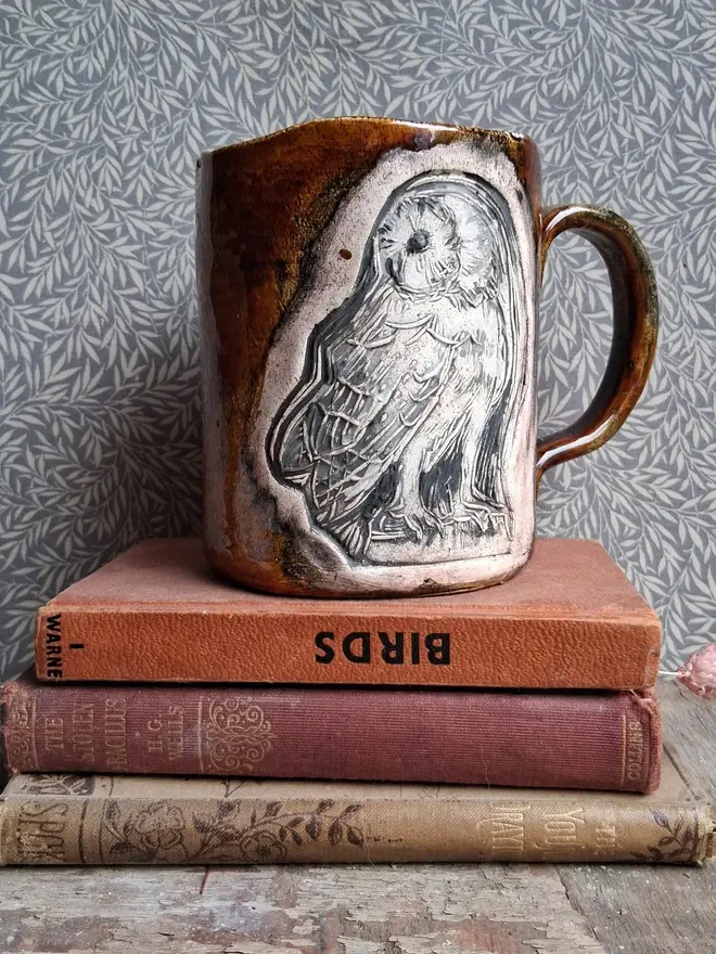 linocut owl ceramic mug. a brown mug bearing an owl design placed on top of a pile of books.