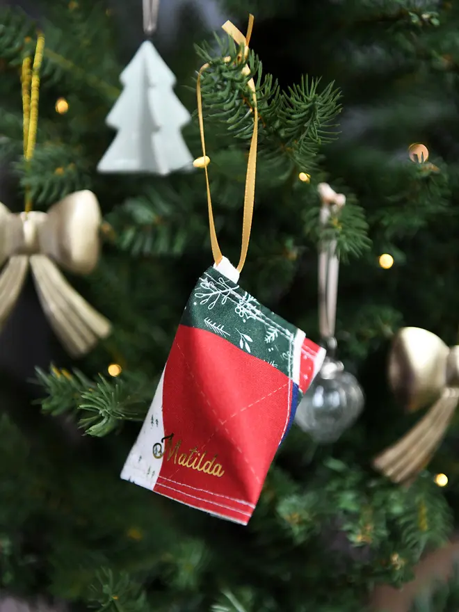 A handmade fabric fortune cookie Christmas decoration in green festive patchwork print, personalised with the name “Matilda” in gold, hangs from a golden ribbon on a Christmas tree branch.