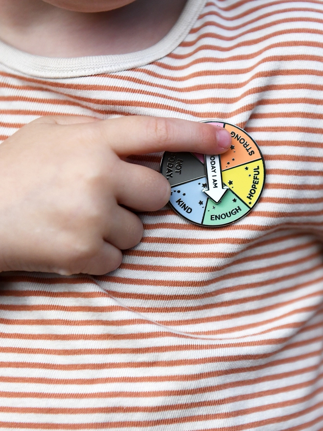 A round enamel pin that looks like a pie chart with six segments, each with positive word, and a white arrow that reads "Today I Am", is pinned to a beige striped top. A child's hand is spinning the arrow,