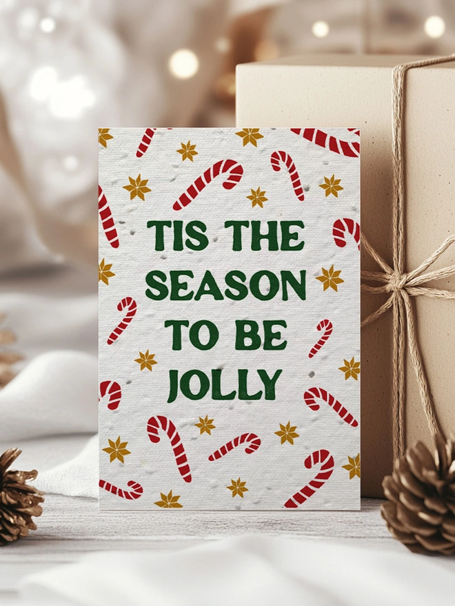Christmas Card with candy canes and gold star pattern with 'Tis the season to be Jolly' in bold forest green text. The card is surrounded by Kraft paper wrapped presents and pine cones.
