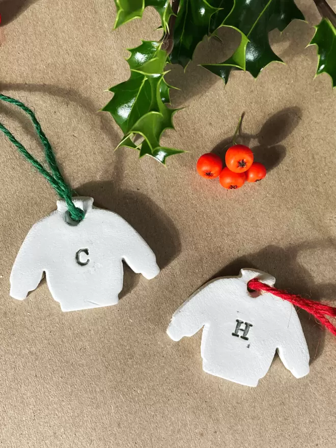 Two clay jumper tags, one with a 'C' and the other a 'H' in the centre of the jumper. One with green twine, the other with red twine. There is a sprig of holly in the background.