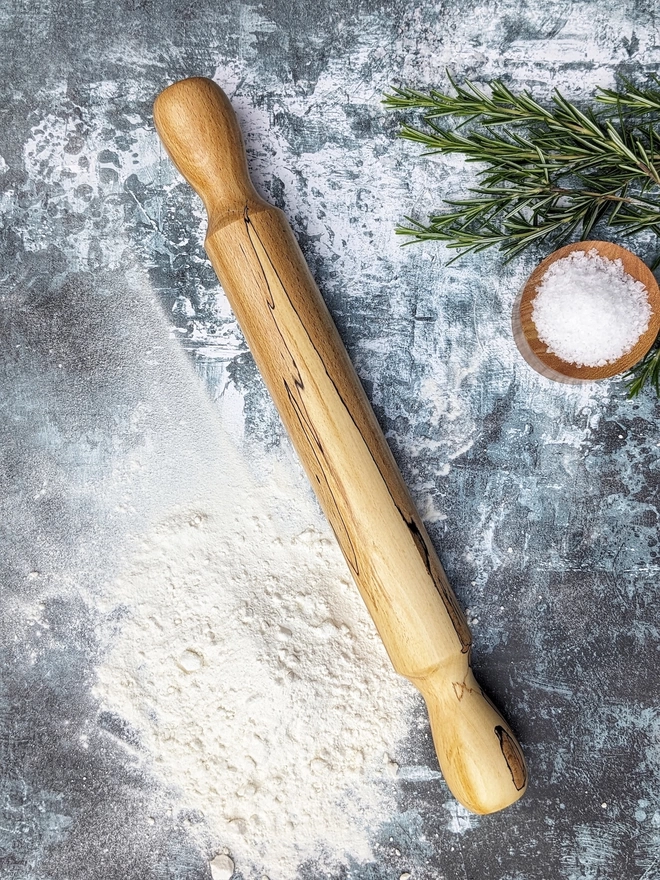 A stunning hand made rolling pin in Spalted Beech by Something From The Turnery. The rolling pin sits on an art house kitchen top amongst flour, rosemary and a wooden salt pot, prepped to bake some delicious bread! Made from Spalted Beech, the rolling pin features two traditional shaped handles amongst it’s own , never repeated natural grain patterns, travelling through golden browns, tans and stunning black lines to outline the changes in the timber. 