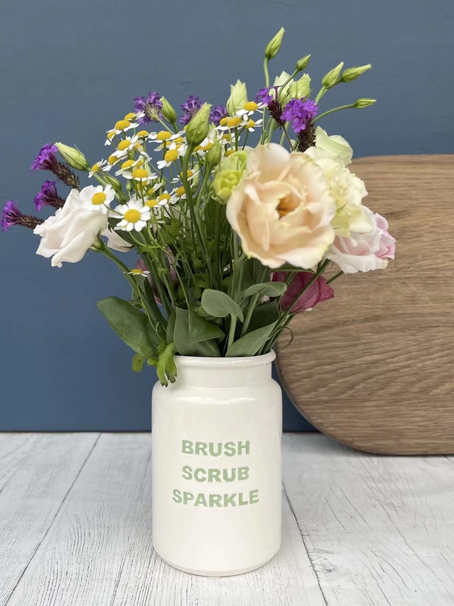 A handmade ceramic ‘Brush Scrub Sparkle’ Pot (lettering in Green) is being use to display flowers.