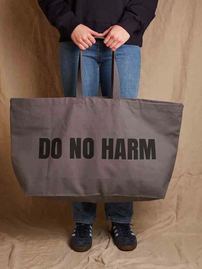 Model holding a massive gunmetal grey cotton canvas tote bag with the words Do No Harm printed in black 
