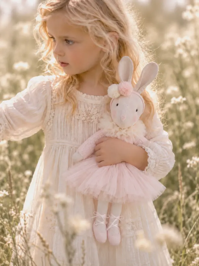 Young girl in an ivory vintage-style dress holding the Verity Isabelle ballerina bunny in a sunlit meadow.