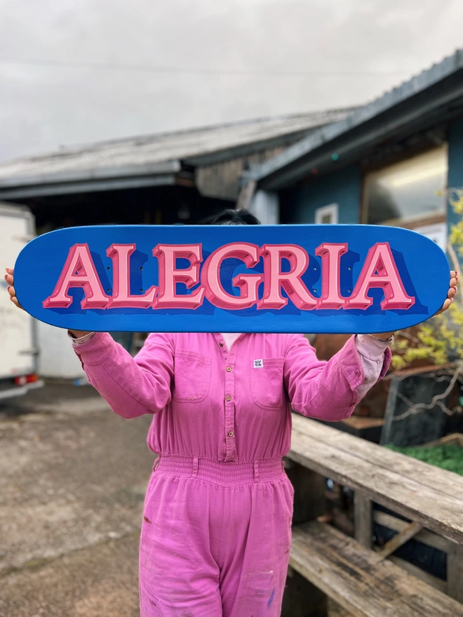 Blue skate deck with 'ALEGRIA' in pastel pink with bright pink outline and shade.