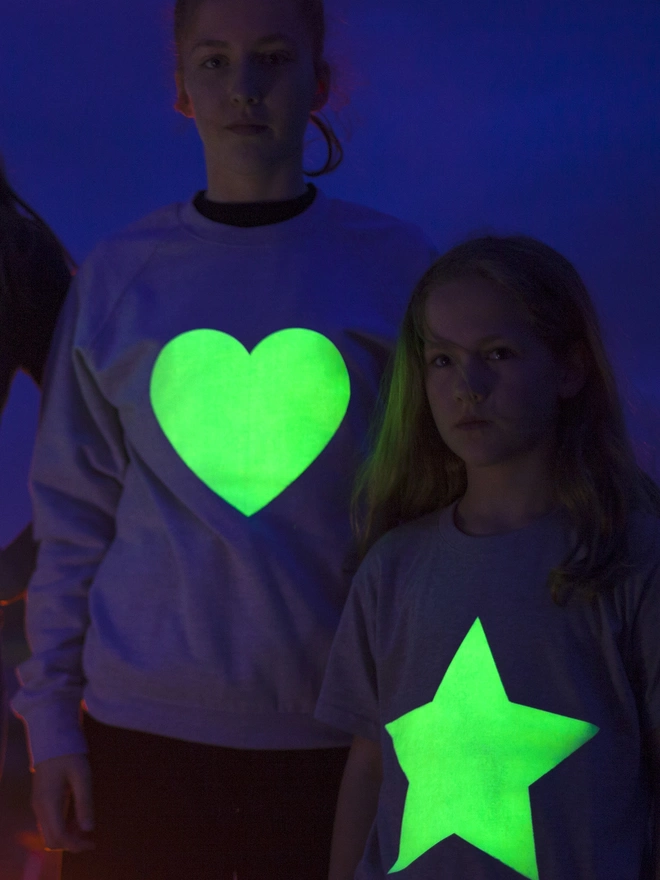 Two girls standing in the dark with glow up sweats on, one with a heart and one with a star