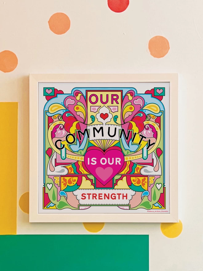 Our Community is Our Strength is written over this bold, symmetrical illustration of yellows, greens and pinks. It is hung in a white square frame on a white wall with yellow, orange, green and blue spots, and a green and yellow rectangle painted in the bottom left hand corner. 