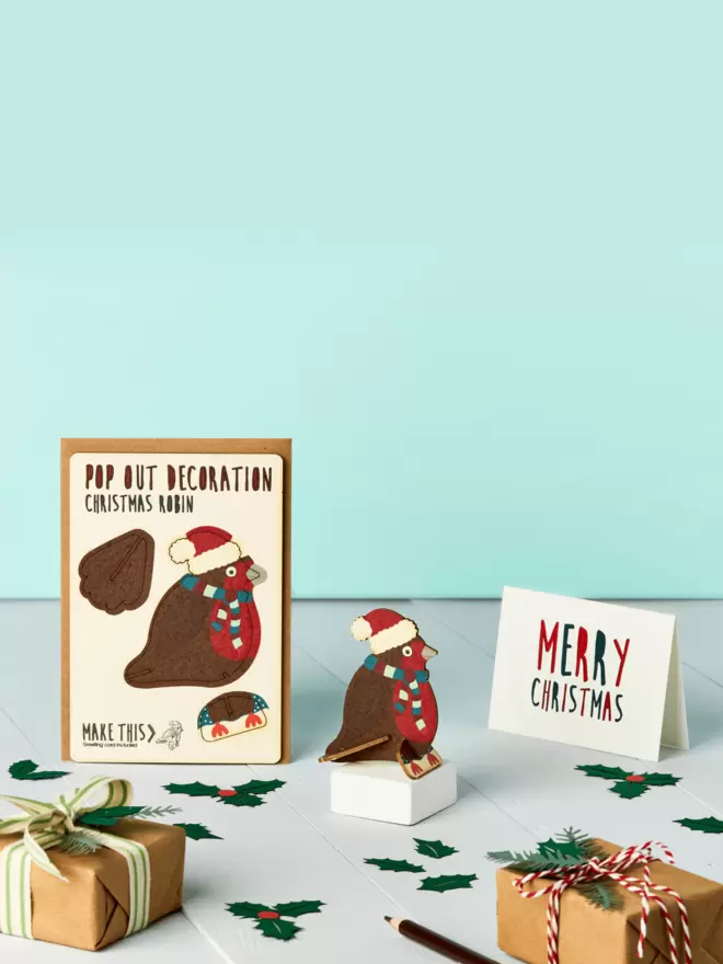 3D laser-cut robin Christmas decoration and Merry Christmas card and brown kraft envelope on top of a wooden desk in front of a pale blue coloured background