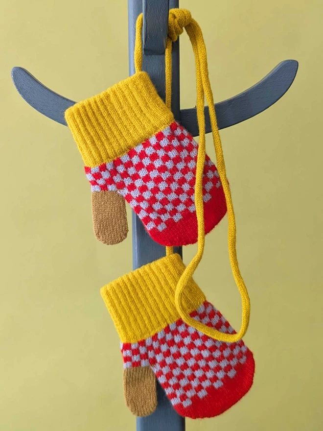 Kids Red And Light Blue Check Mittens On A String Hanging On A Coat Stand In Front Of  A Green Background
