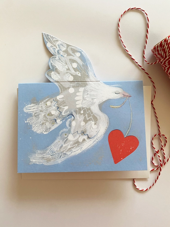 Esther Kent greetings card showing white flying dove in a light blue background, carrying a vibrant red heart on a string