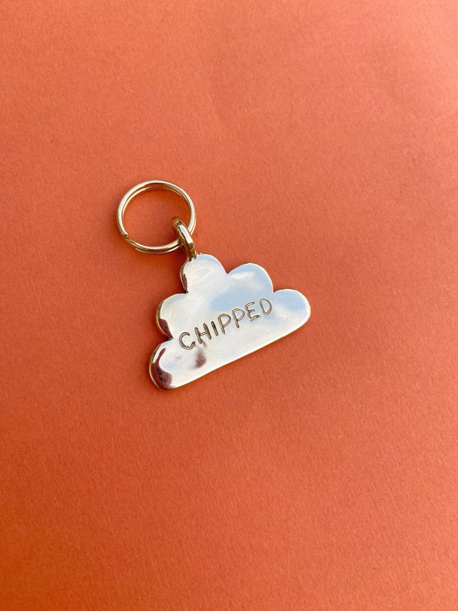 the back of a brass pet tag in the shape of a cloud, with 'chipped' engraved into it, on a burnt orange background.
