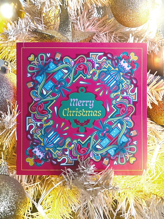 An abstract, square Christmas card design, with Merry Christmas in the centre, surrounded by a multi-coloured design including crackers and presents, on a red background, rests in white and gold tinsel and baubles.