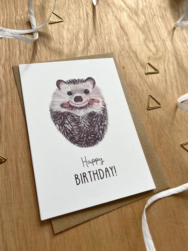 A greetings card with a white background featuring a baby hedgehog on their back with the phrase "happy birthday" underneath