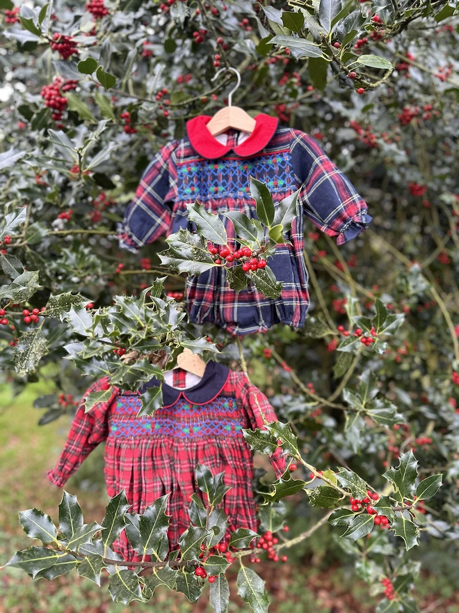 Two christmas rompers in red tartan and navy tartan with contrasting collars hang in the holly.