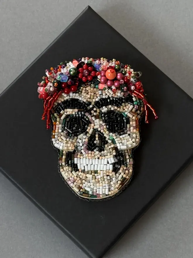 Frida Kahlo Skull Beaded Brooch displayed against a black box, on a grey table. 