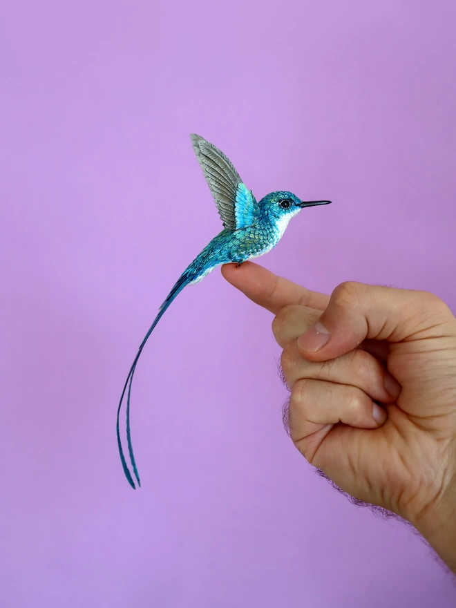 paper hummingbird balancing on finger in front of purple background
