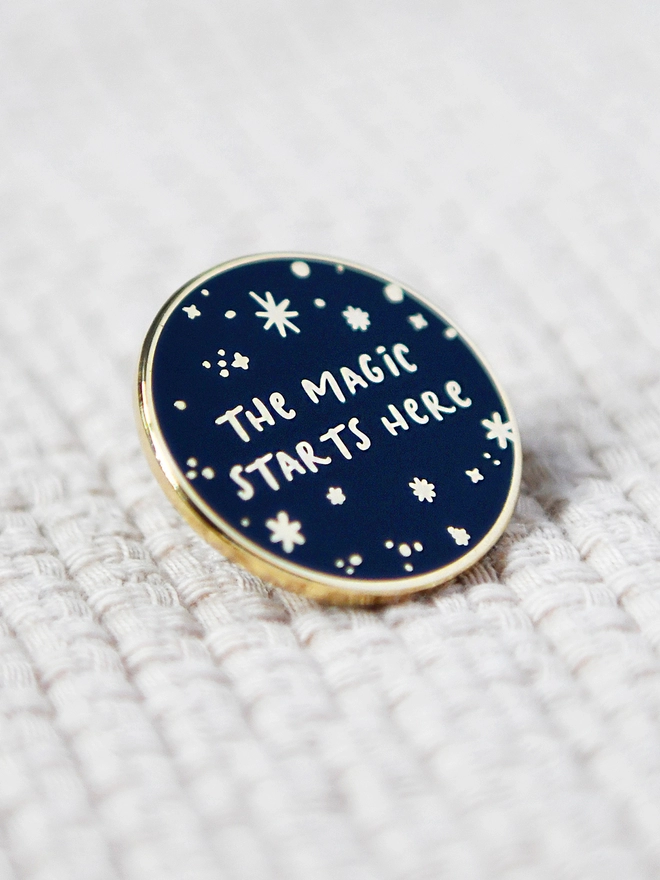 A navy blue and gold enamel pin badges with a starry design and the words "The magic starts here" rests on white fabric.