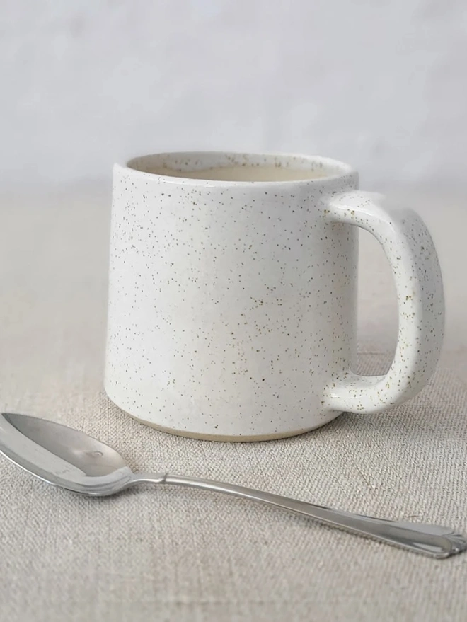 Ceramic Standard Mug, a colourful ceramic mug sitting on a plain surface against a grey backdrop. It is accompanied by a spoon. 