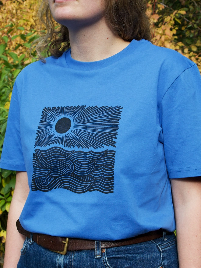 woman wearing a blue t-shirt with a black handprinted design on the front depicting the sun and the sea