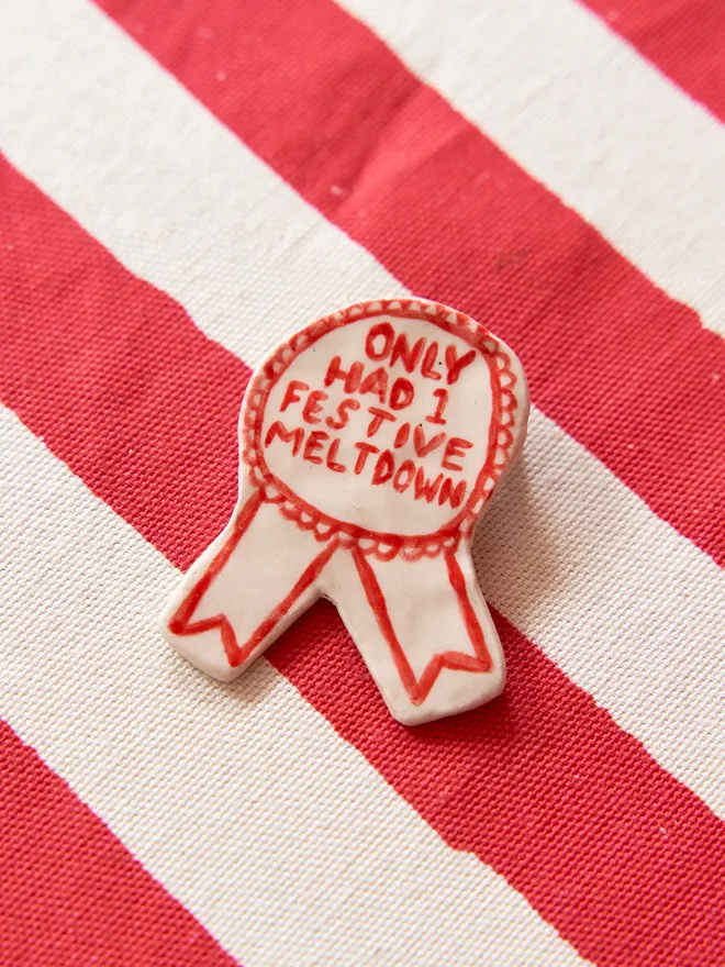 'Only had 1 Festive Meltdown' hand-painted on a ceramic Christmas award badge in red, atop a striped red and white tablecloth