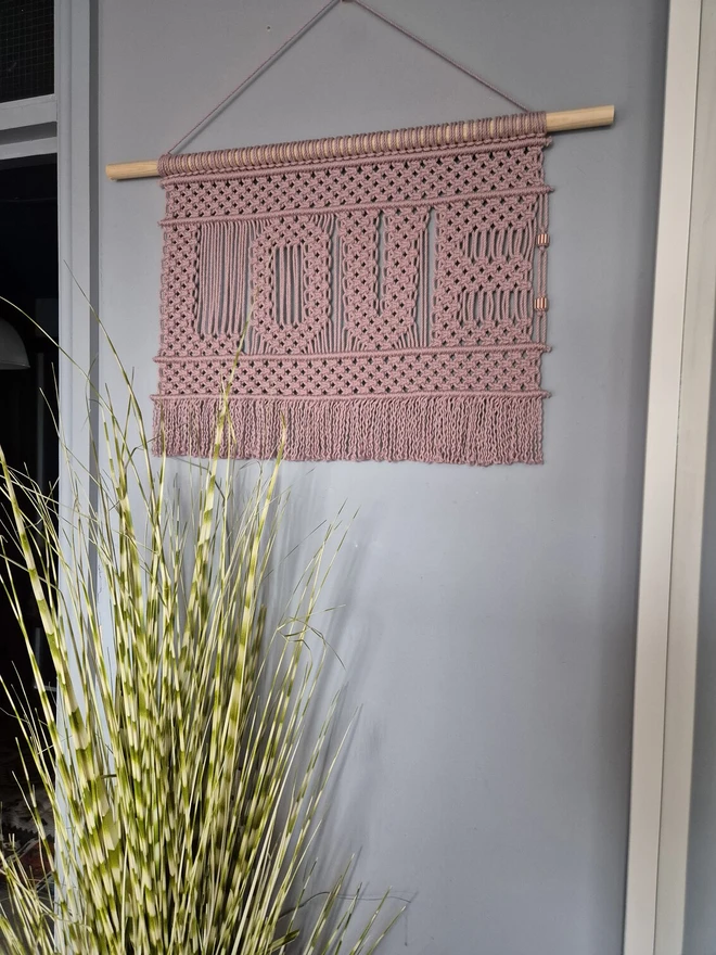 handcrafted macrame wall hanging with the word 'Love' intricately woven in pink rope, featuring detailed classic macrame knots, fringed edges and a wooden dowel for display