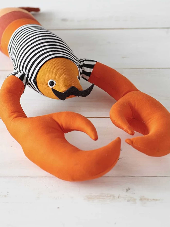 Orange fabric toy lobster wearing black and white t shirt with a black moustache lying on white floorboards 