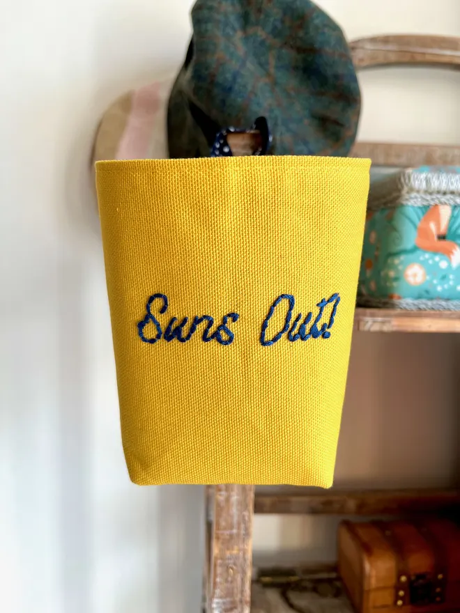 Yellow storage bag hanging on a wooden stand, the front embroidered with ‘Suns Out’ in navy thread.