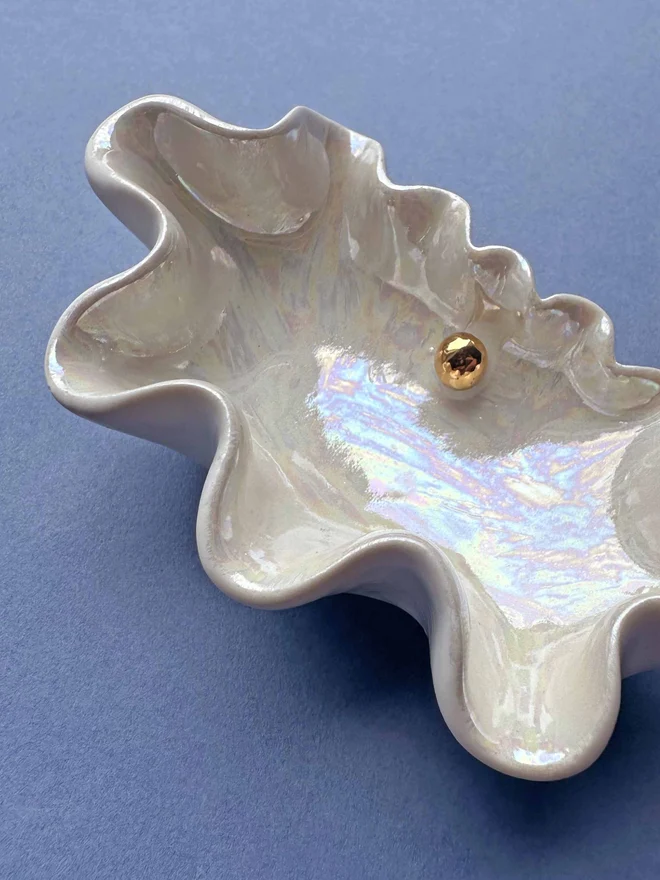 Porcelain clam shell pinch pot in shimmery white with gold mother of pearl in the middle of the shell, on a blue background