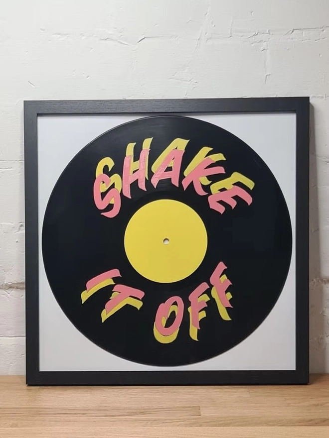 'Shake It Off' 12' Vinyl Artwork, a colourful vinyl sitting in a black frame on a wooden surface against a stone backdrop. 