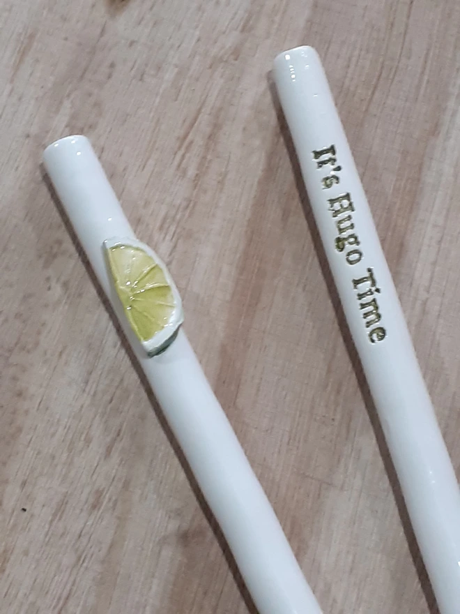 Photo of two handmade ceramic drinking straws on a wooden surface — one with a sculpted lime slice motif and the other with the phrase “It’s Hugo Time” in glossy lettering, inspired by the classic Hugo Spritz cocktail.