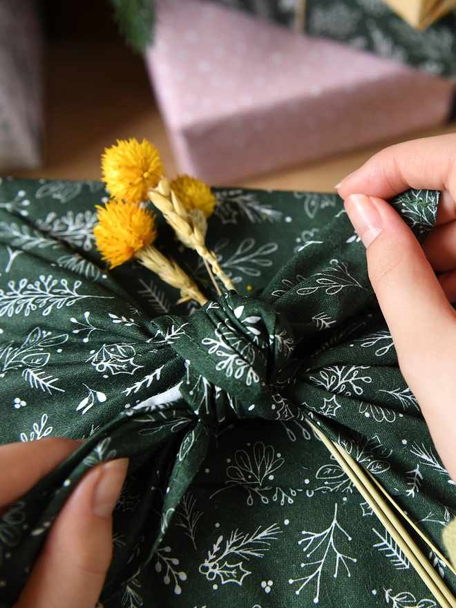A gift is being wrapped in deep green cotton fabric wrap with a botanical design, and a small posy of yellow flowers is being tucked into the knot.