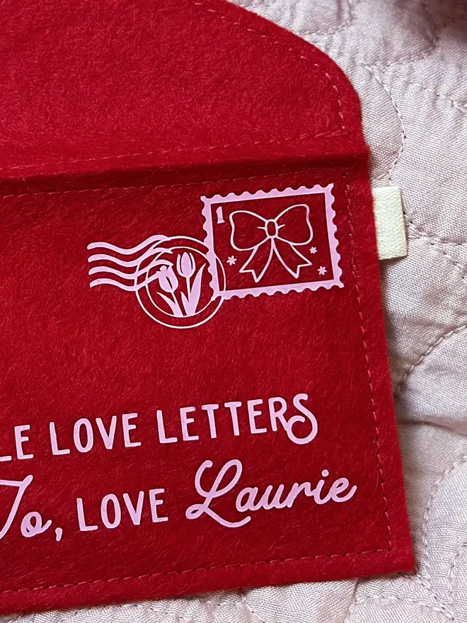 A red felt envelope with a pale pink stamp and postmark on. The stamp had a bow design and the postmark has two tulips inside.