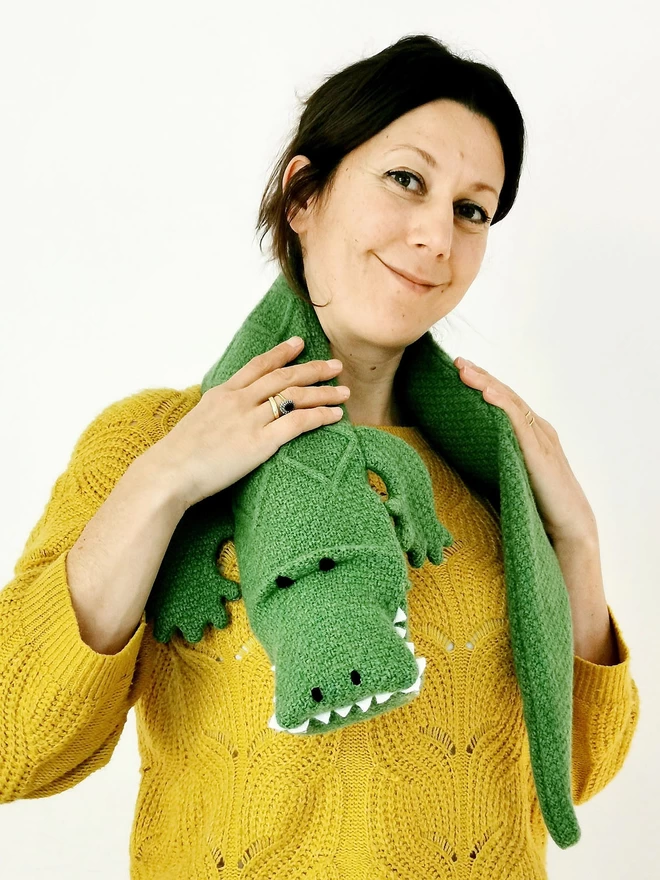 A smiling woman stands facing the camera with a bright green knitted crocodile hot water bottle draped around her neck.