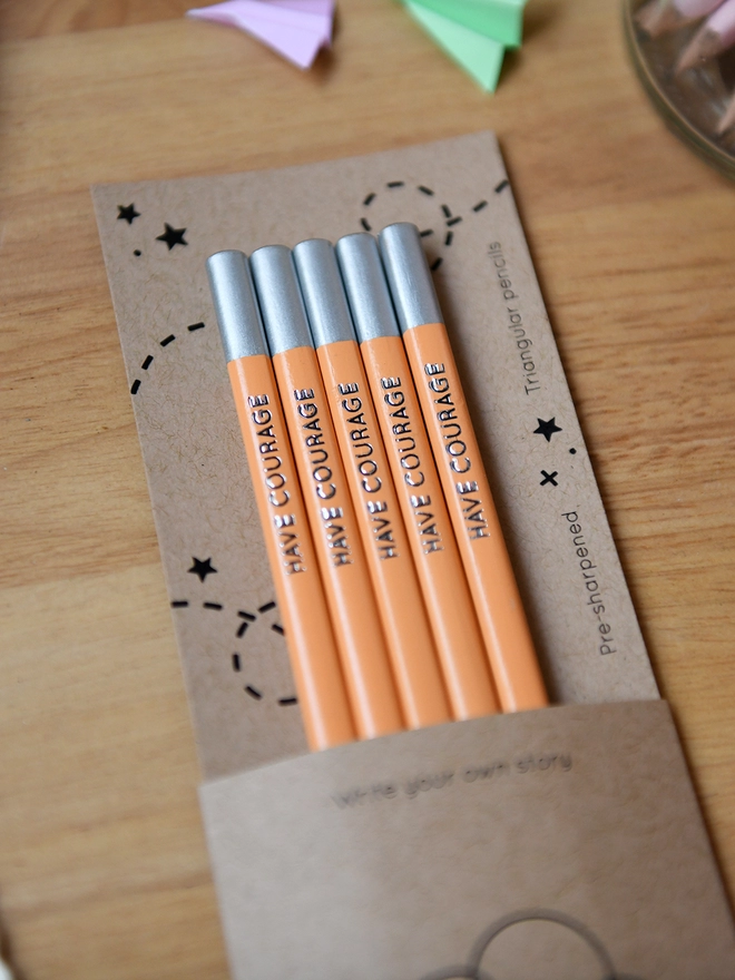 Five orange pencils with the words Have Courage along the side of each one, are tucked into cardboard packaging on a wooden desk.
