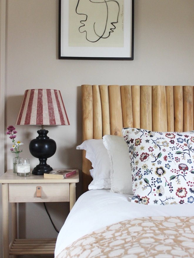 Empire lampshade with bold burgundy and pink stripes, styled on a black table lamp beside a bed with a wooden Tikamoon headboard. A colourful lampshade and classic striped lampshade for bedrooms.