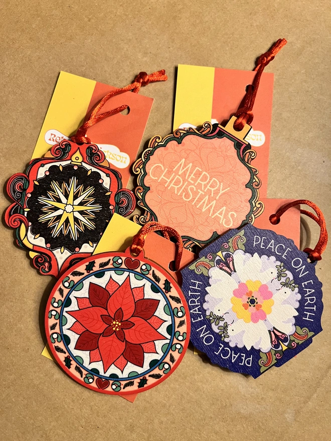 Multi coloured wooden tree decorations in a group of 4. They are tied with red ribbons and are lying on a brown card background with business cards with Rebecca Strickson on them. 