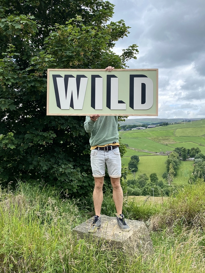 A person surrounded by moorland holding a large Woden sign reading WILD on a soft green painted back board. 
