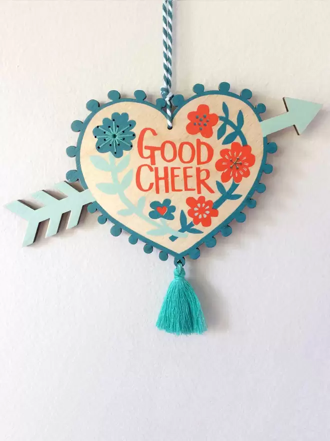 'Good Cheer' is hand printed on a floral wooden love heart charm, with an arrow piercing it