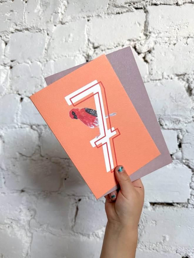 bright orange card with a big white number 7 and an illustrated madagascan fody bird with a purple envelope