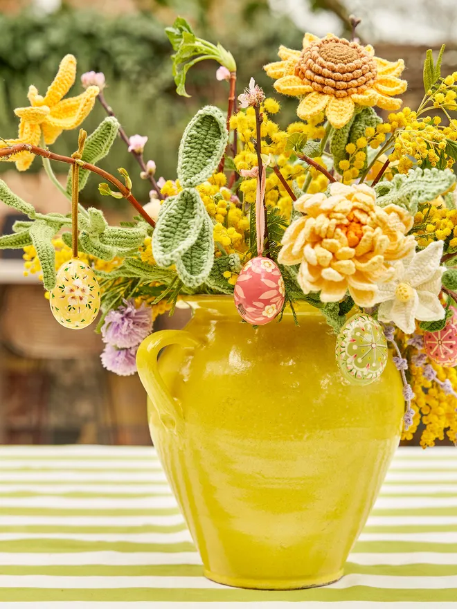 Assorted crochet flowers assorted in a bright yellow vase on a green and white striped tablecloth in a garden setting, with several wooden painted hanging eggs