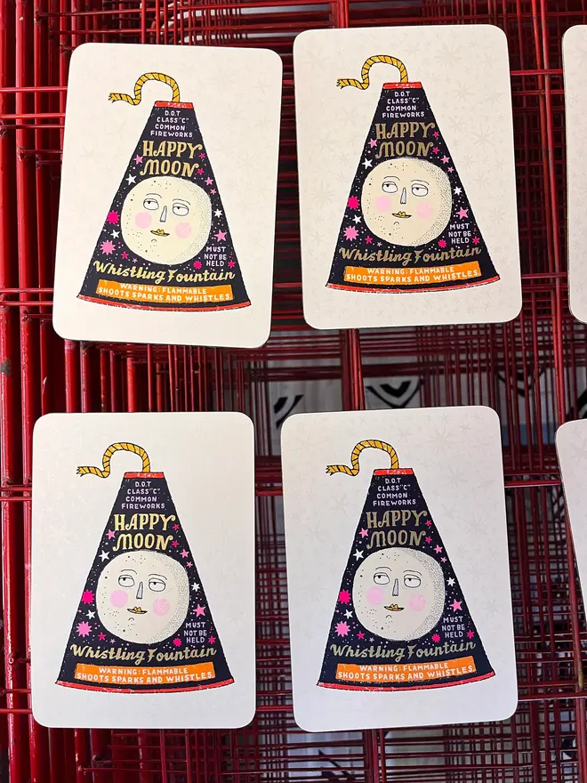 A Flash Of Brilliance Moon Firework Screenprint, a colourful firework design in the centre of a white screenprint. It is placed on a studio rack joined by other screenprints. 